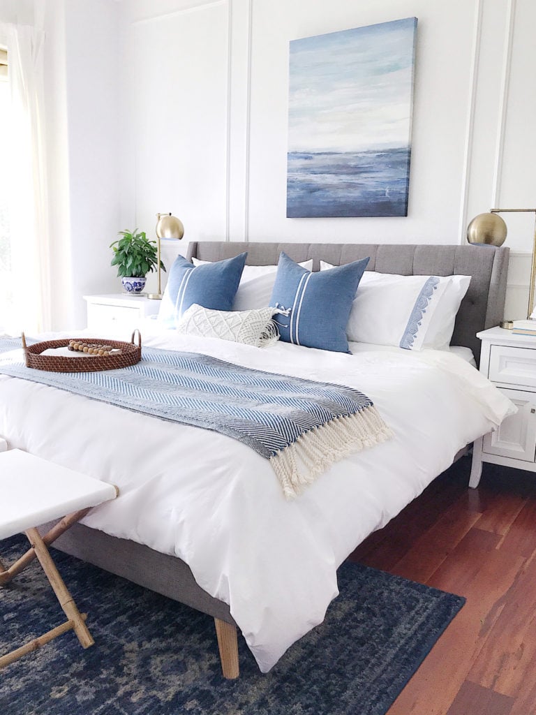 Master bedroom with white bedding, a blue rug, stools, blue and white pillows, modern coastal artwork, a gray upholstered bed, and picture frame wall molding - jane at home