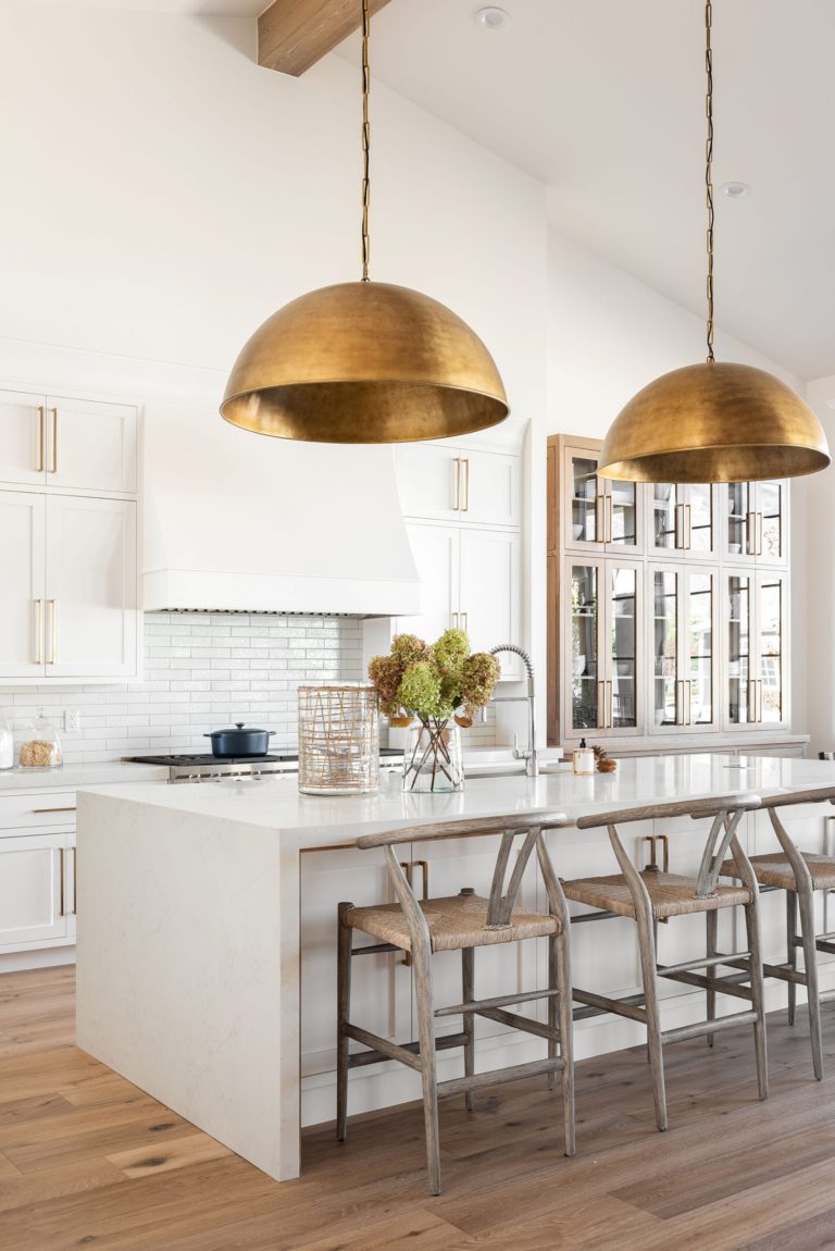 Beautiful modern open kitchen design with white cabinets, brass and gold accents, large island with waterfall countertop, wood beams and large brass pendant lights over island - Studio McGee Sunset House Project