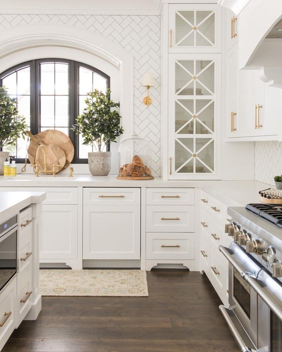 Love this beautiful timeless kitchen design with herringbone tile backsplash, arched window, white cabinets, and brass hardware and finishes