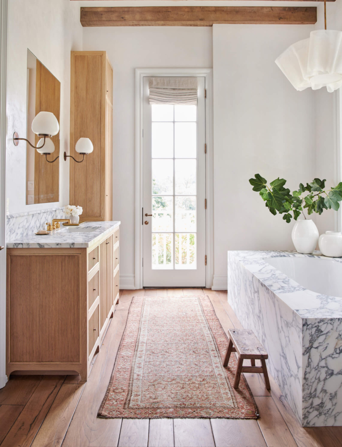 Love this beautiful and elegant bathroom design with a marble bathtub, warm wood vanity and linen cabinets, wood beams, vintage rug, brass faucets, and modern chandelier - amber interiors