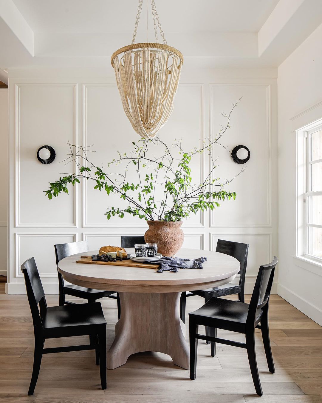 A beautiful modern dining room decorated with fresh branches, rustic pottery, a wood charcuterie board, a round dining table in light wood, and modern black side chairs - kelsey leigh design co.