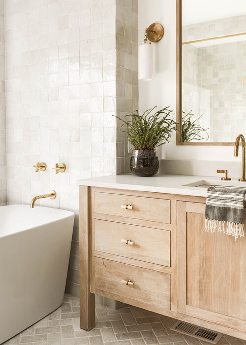 Love this beautiful small bathroom design with a light oak wood vanity cabinet, zellige tile tub surround, and brass finishes on the faucets, mirror, lighting, and hardware - henri design