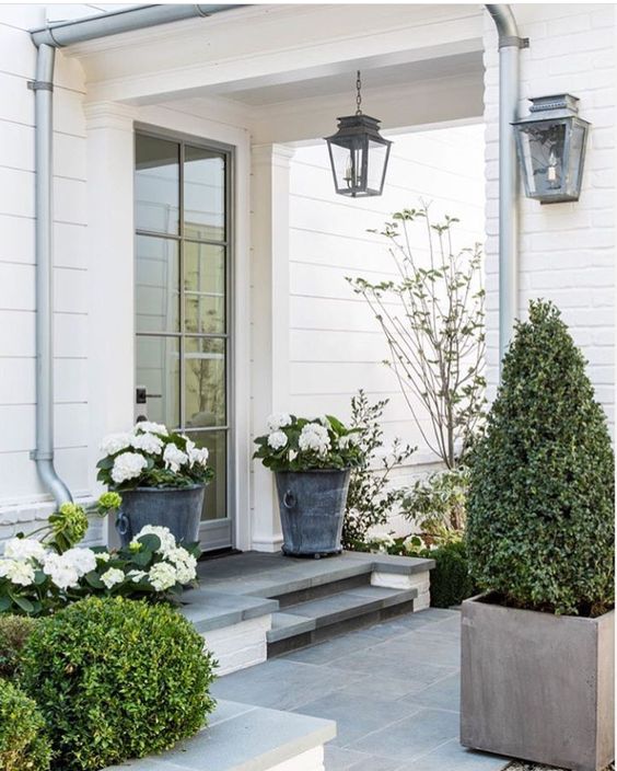 Love this beautiful front porch with potted boxwoods, white flowers, and container plants, with black lantern outdoor lighting, from Gianetti Home